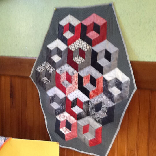 Photo d'un travail de couture en patchwork, accroché sur un mur. Il représente des formes hexagonales dans les tons gris et rouge.